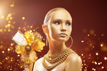 Festive golden jewels, luxury style. A gorgeous girl with golden skin and hair wearing golden jewelry and holding a golden flower. Magical golden lights shine around. Holiday luxury beauty concept.の写真素材