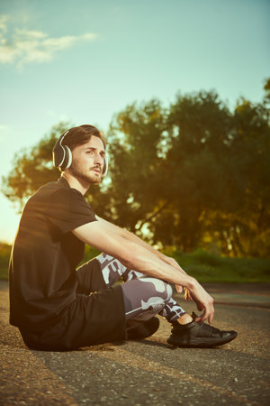 Sporty lifestyle. An active guy in sportswear relaxes on the ground in the park after training listening to music on his headphones.の写真素材