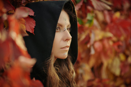 A beautiful shaman woman with long curly hair and ethnic ornaments on her face stands among red foliage in the forest. Fantasy. Ethnic culture. Shaman woman.の写真素材