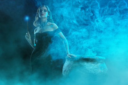 A beautiful young witch in an elegant black off-the-shoulder dress conjures at night over a cauldron with a magic wand. Halloween magic. Copy space.の写真素材
