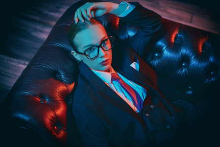Beautiful confident lady poses in a stylish pantsuit with a tie and glasses on a leather sofa....