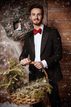 A handsome young man in elegant suit standing by a snow-covered wooden house with a basket and christmas-tree branches. Merry Christmas and Happy New Year!の写真素材
