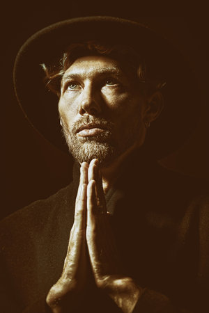 A calm handsome mature man with beard and moustache in black coat and black hat standing with hands clasped in prayer and looking up. Black background. Art portrait. Male beauty.の写真素材