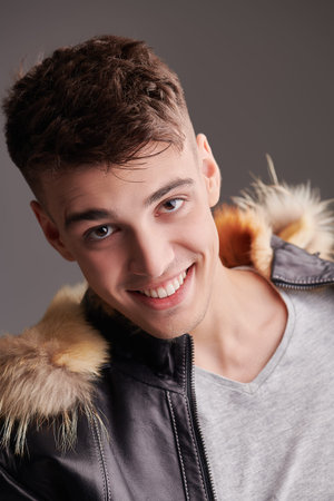 Close up shot of a happy handsome brunet man posing in a winter jacket with fur hood on a grey background. Men's beauty, winter fashion. Emotions.の写真素材