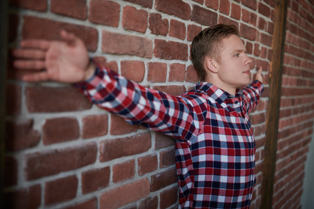Young people, lifestyle. A handsome modern guy in a chekered shirt stands in a loft style room by a brick wall. Casual male fashion.の写真素材