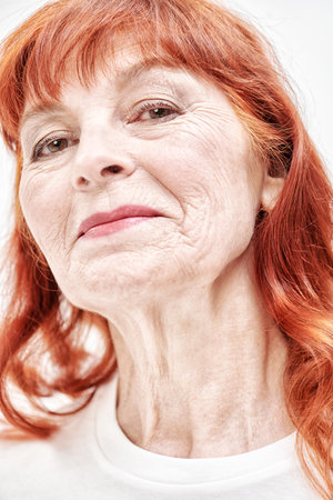 A close-up portrait of a beautiful elderly woman with bright red hair wearing a white blouse. White background. Senior people concept. Make-up and cosmetics for the elderly.の写真素材