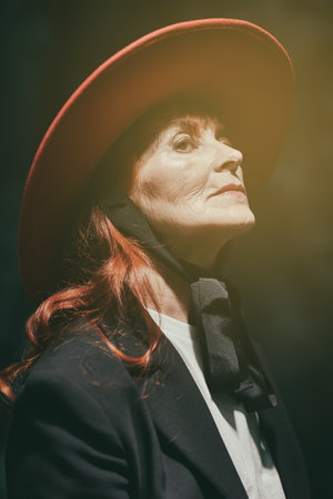 An elegant senior woman with red hair posing in a stylish suit and a red hat in the studio on a black background. Fashion and beauty for the elderly.の写真素材