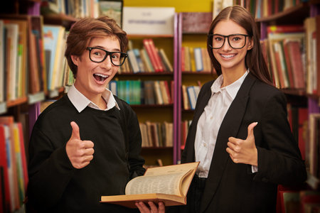 Two happy students in glasses and in black and white uniform stand in a library choosing a book....