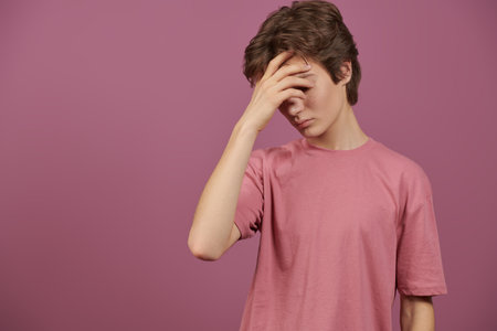 Adolescence, emotions and experiences. Portrait of a teenage boy who stands sadly covering his face with his hand. Studio background with copy space.の写真素材