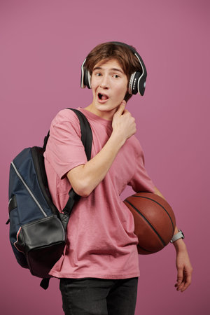 A modern teenager boy in headphones with a school backpack behind his back and a basketball makes a...