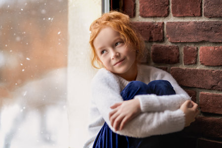 A portrait of a cute red-haired little girl in casual clothes sitting thoughtfully by the window. Natural lighting. Children' fashion. Childhood and emotions. Winter snowy day.の写真素材