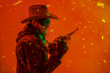 Studio portrait of a man dressed in the style of the Wild West fighting with his gun during apocalyptic time. Post apocalyptic cowboy. Red background with burning pieces after explosion.の写真素材