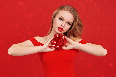 Elegant blonde girl with bright evening make-up in a gorgeous red dress holds a festive red gift bow. Festive red studio background. Make-up and cosmetics. Holiday celebration.の写真素材