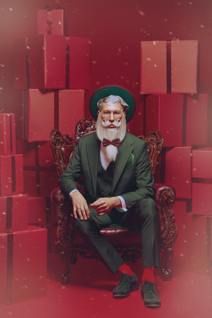 Christmas, New Year and Fashion. An elegant Santa Claus with a beautiful festive beard and mustache sits in an armchair in a stylish green suit. Festive red background with many gifts.の写真素材