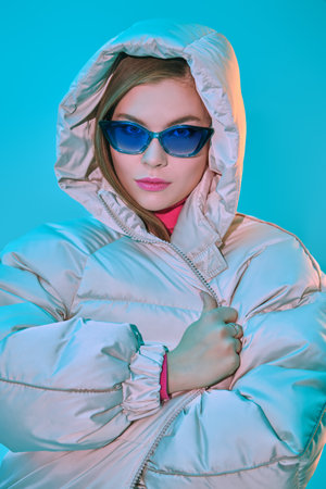 Portrait of a beautiful fashion model girl posing in a white down jacket and elegant blue glasses on a blue studio background. Winter fashion.の写真素材