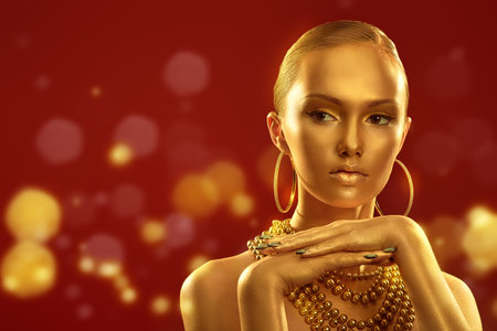 Gorgeous young woman with golden skin, makeup and hair posing in golden jewellery on a festive red background with sparkling golden lights. Christmas beauty girl. Gold luxury concept. Copy space.の写真素材