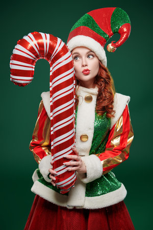 Funny pretty Christmas fairy elf girl in a festive costume holds big candy cane and flirts on a...