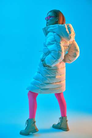 Full length portrait of a fashion model girl posing in a long white down jacket and bright fuchsia glasses and tights. Blue studio background. New winter collection of clothes and accessories.の写真素材