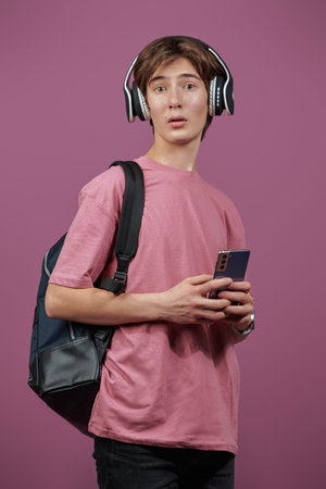 A surprised modern teenager boy in headphones with a school backpack behind his back and a...