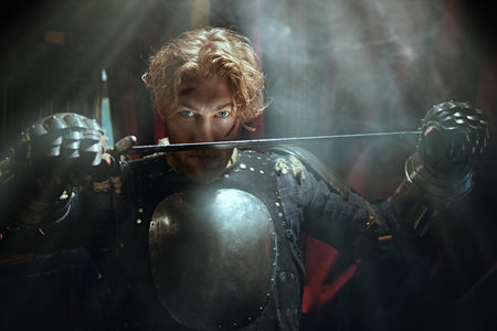A scene from a chivalric romance. A handsome noble medieval knight in armor stands with a raised...