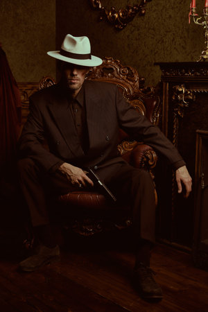A tough gangster man in an elegant black suit and a white hat sits in a chair with a gun in his hand in a luxury apartment. Retro style. The criminal world of the mafia. Detective movie.の写真素材