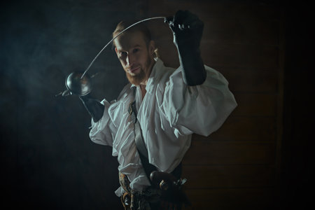Portrait of a handsome brave musketeer in a white shirt standing in a dark vintage room and holding a rapier in his hands. 16-17 centuries. Historical novel. The era of romanticism.の写真素材