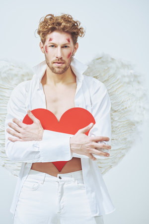 Handsome courageous male Cupid angel with wounds on his face presses a red heart to himself, standing in white clothes and with white wings on a white background. God of love Cupid. Valentine's Day.の写真素材