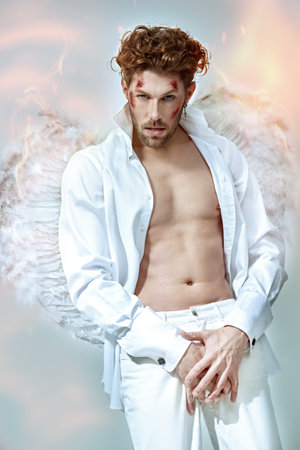 Fantasy hero lover. Attractive male Cupid angel with wounds on his face, in white clothes and with white wings looking seductively into the camera. God of love Cupid. Valentine's Day.の写真素材