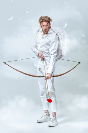 Love concept. Portrait of the warlike handsome God of love Cupid with wounds on his face, going to shoot an arrow at his victim. White background with clouds and feathers around. Valentine's Day.の写真素材