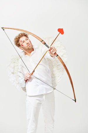 Valentine's Day. A warlike handsome man, the God of love Cupid with wounds on his face, intends to shoot an arrow at his victim. Fantasy. Studio white background.の写真素材