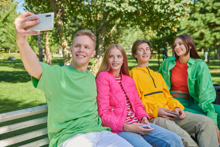 Carefree friends teengers in bright youth clothes sitting in the park on the bench and taking a...