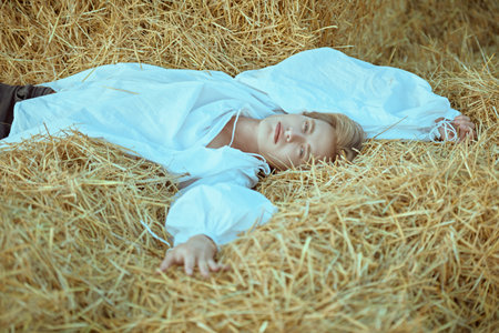 The era of romanticism.  A romantic handsome young man with blond hair in a vintage white shirt with wide sleeves lies relaxed in a haystack in a field. The hero of a love-adventure novel.の写真素材