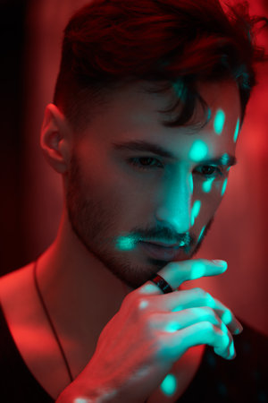 An art shot of a masculine handsome brunette man standing in a dark room with red lighting. Men's emotions and beauty. Beams of light fall on him.の写真素材
