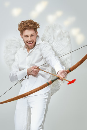 Valentine's Day. A handsome man, the God of love Cupid intends to shoot an arrow at his victim. Fantasy. Studio white background.の写真素材