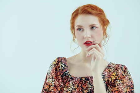 A studio shot of a beautiful ginger girl with delicate fresh make-up posing against a white wall in a floral dress. Spring and summer fashion, beauty. Romantic feminine style. Copy space.の写真素材