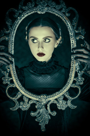 Portrait of a dramatic brunette girl in an elegant black velvet dress and lace gloves, who stands in the night on a dark vintage background with a mirror frame in her hands. Gothic novel, mysticism.の写真素材