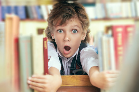 A cute surprised boy stands in a bookshop peeking out from behind the books on the bookshelf. Education and clever children.の写真素材