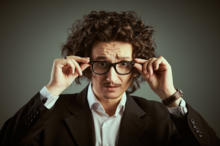 A stylish Italian man with dark curly hair and a small mustache, dressed in a classic black suit, anxiously looks at the camera through his glasses. Studio shot in retro style. Male characters.の写真素材