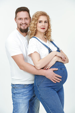 A loving husband and a beautiful pregnant wife are expecting a baby. Loving spouses in white t-shirts and blue jeans hug their tummy with a baby on a white studio background.の写真素材