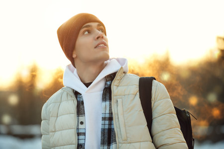 A handsome happy young man in warm clothes stands in a winter park at sunset and admires the beauty of nature.の写真素材
