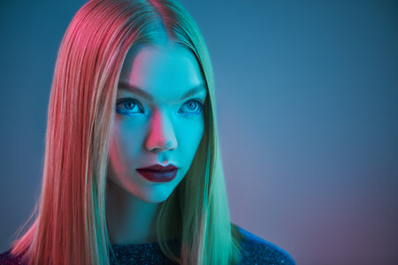 Stunning blonde girl with shiny glitter makeup and a sparkling evening dress poses on a blue background in mixed colored light and copy space. Glitter, fashion and style. Hairstyle.の写真素材