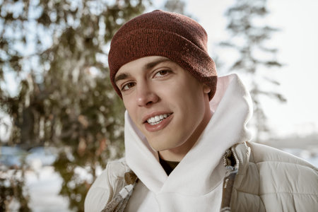 Handsome happy young man in a knitted hat and down jacket stands in a winter park and smiles. Happy...