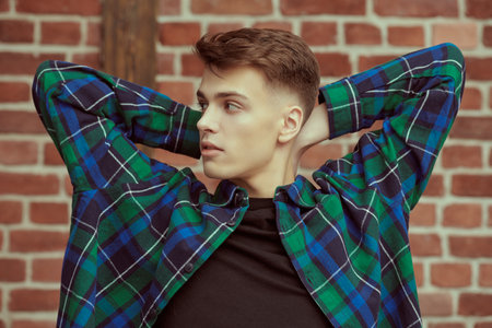 A handsome young man in a flannel plaid shirt sits relaxed against a brick wall and looks to the side. Youth fashion. Studio portrait.の写真素材