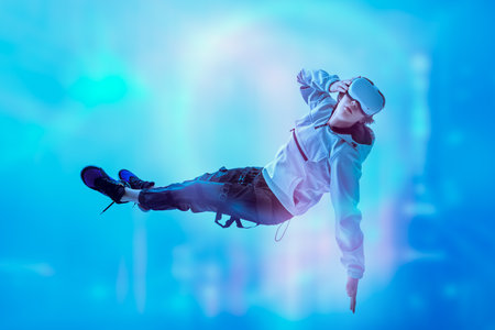 A modern teenage boy flies through the air wearing virtual reality glasses, immersing himself in the game. Virtual reality games. Sky blue background with a glowing energy field.の写真素材