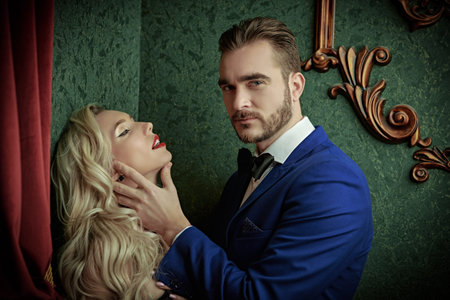 A handsome confident man in an elegant suit imperiously hugs a gorgeous sexy blonde while standing...
