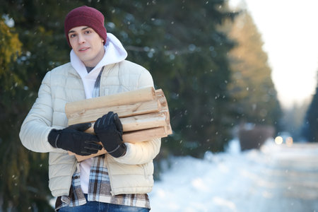 A young man in warm winter clothes holds an armful of firewood, standing in a winter snowy forest, and smiles joyfully. Active recreation in winter, travel and tourism.の写真素材
