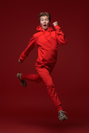 Excited teen boy in a red tracksuit jumps with joy. Happiness, joyful emotions, success. Full-length studio portrait against a red background.の写真素材