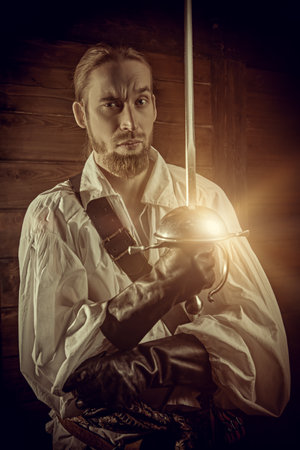 A brave musketeer in a white shirt fights with rapier in a wooden house. 16-17th centuries. Historical novel. The era of romanticism.の写真素材