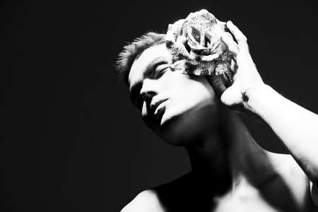 Art black-and-white portrait of a sensual handsome young man with black smokey eyes posing with naked body and rose on a black studio background. Mixed light. Male beauty concept.の写真素材