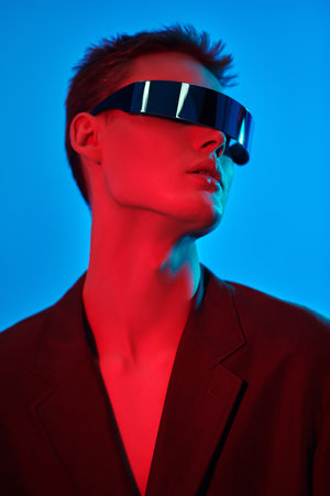 Portrait of a stylish young man posing in mirrored futuristic glasses in mixed neon blue and crimson light. Blue studio background. Glasses style. Fashion concept.の写真素材